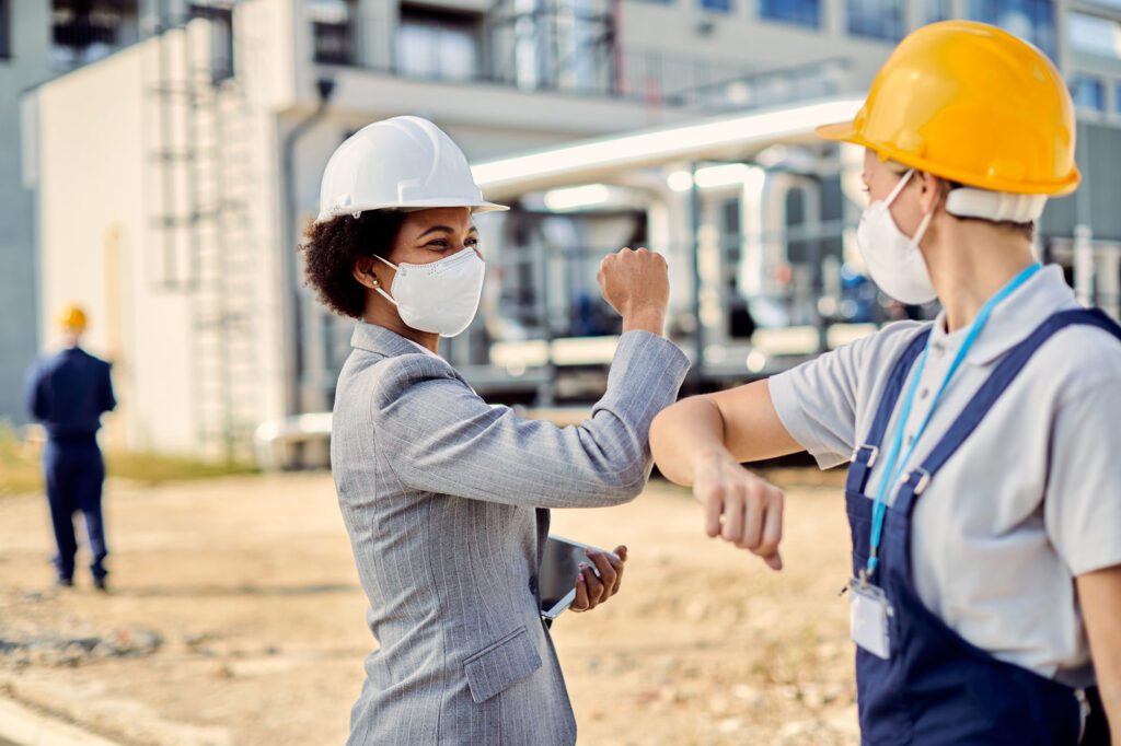 African American civil engineer and worker wearing face masks while elbow bumping at construction site.