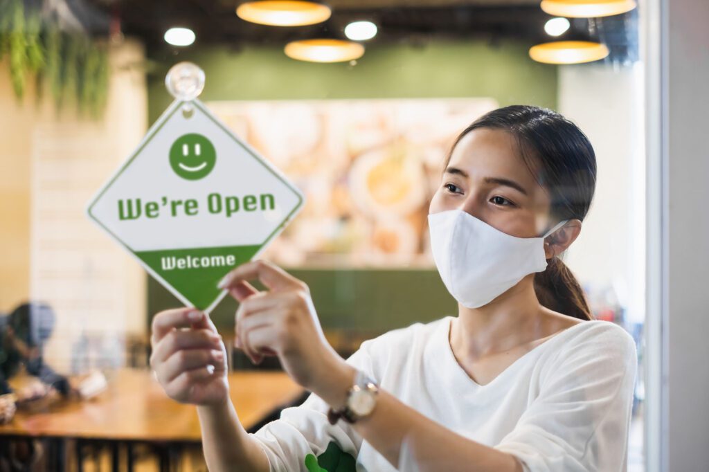 Business owner Asian woman wear protective face mask ppe hanging open sign at her restaurant / café, open again after lock down due to outbreak of coronavirus covid-19