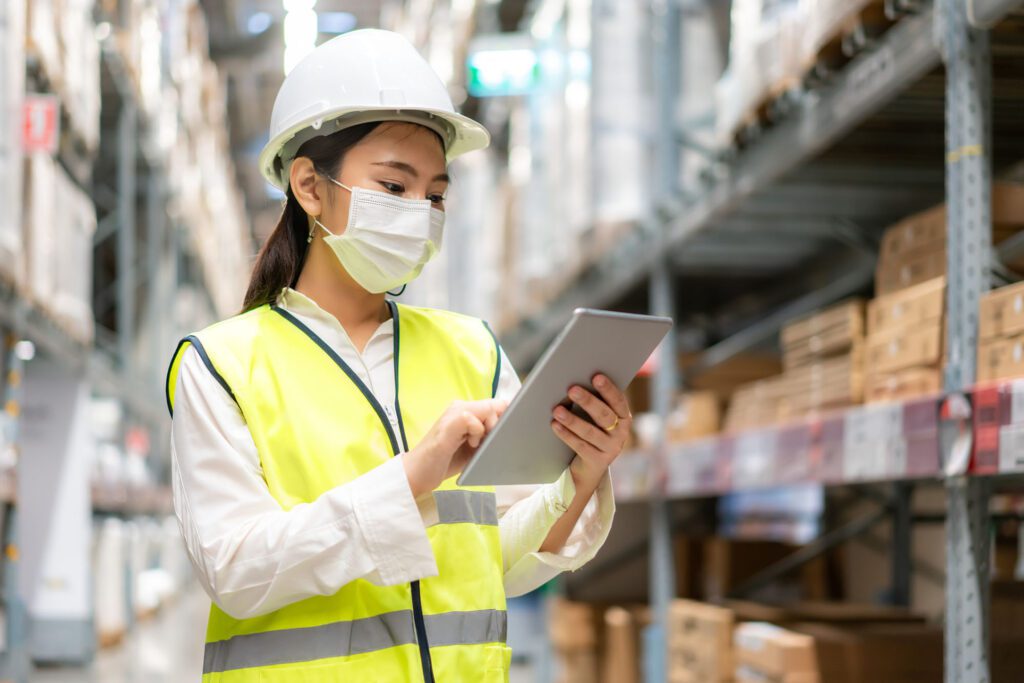 Young asian woman auditor or trainee staff wears mask working during the COVID pandemic in store warehouse shipping industrial. looking up and checks the number of items store by digital tablet.