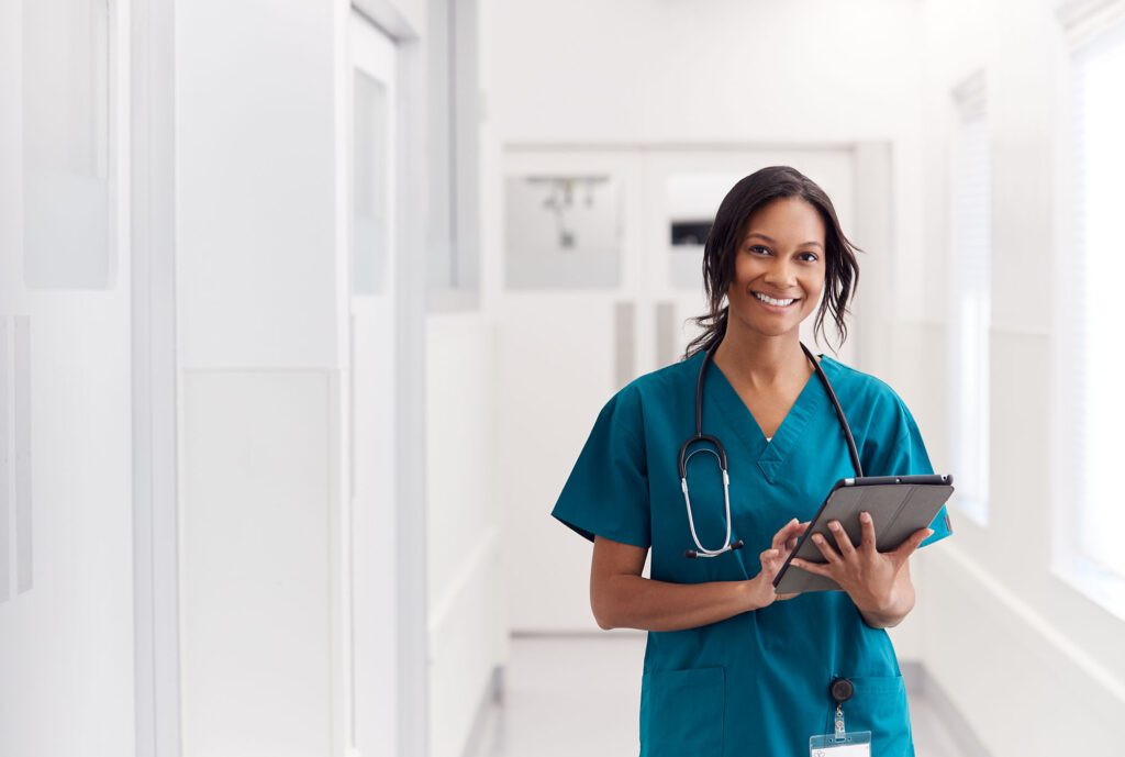 Portrait Of Smiling Female Doctor Wearing Scrubs In Hospital Corridor Holding Digital Tablet