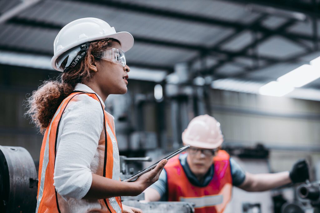 Female industrial engineer wearing a white helmet while standing in a heavy industrial factory behind she talking with workers, Various metal parts of the project