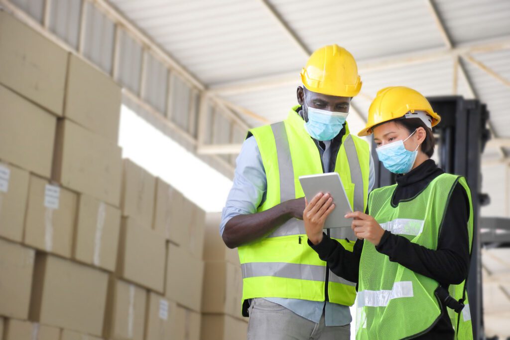 African American and Asian workers wearing facial mask and safety vest working in warehouse checking for the inventory using digital tablet during new normal after covid