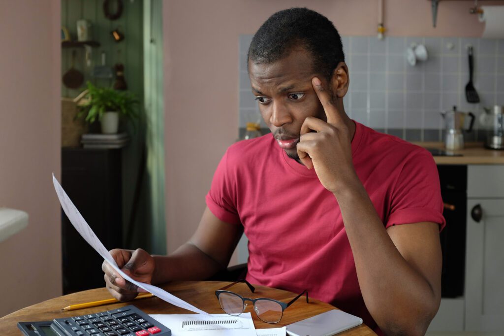 Horizontal shot of handsome African American man sitting at table at home trying to figure out his financial situation analyzing documents, bills, looking nervous and worried because of problems